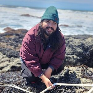 Chris Hernandez kneeling on a rocky intertidal shore with the ocean in the background. He is smiling and holding a transect.
