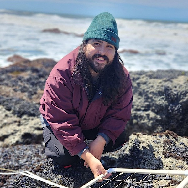 Chris Hernandez kneeling on a rocky intertidal shore with the ocean in the background. He is smiling and holding a transect.