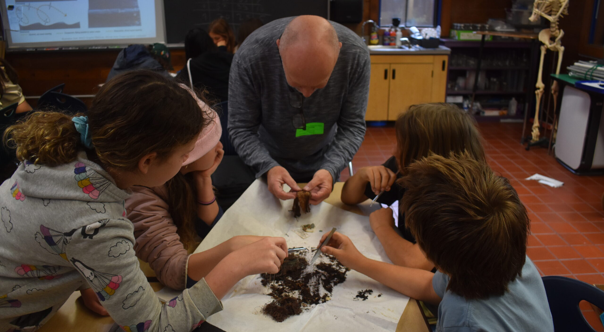 Pete Winch and students huddle around a table with vegetation and dirt. Pete is showing students how to sort through the dirt to find squid beaks.