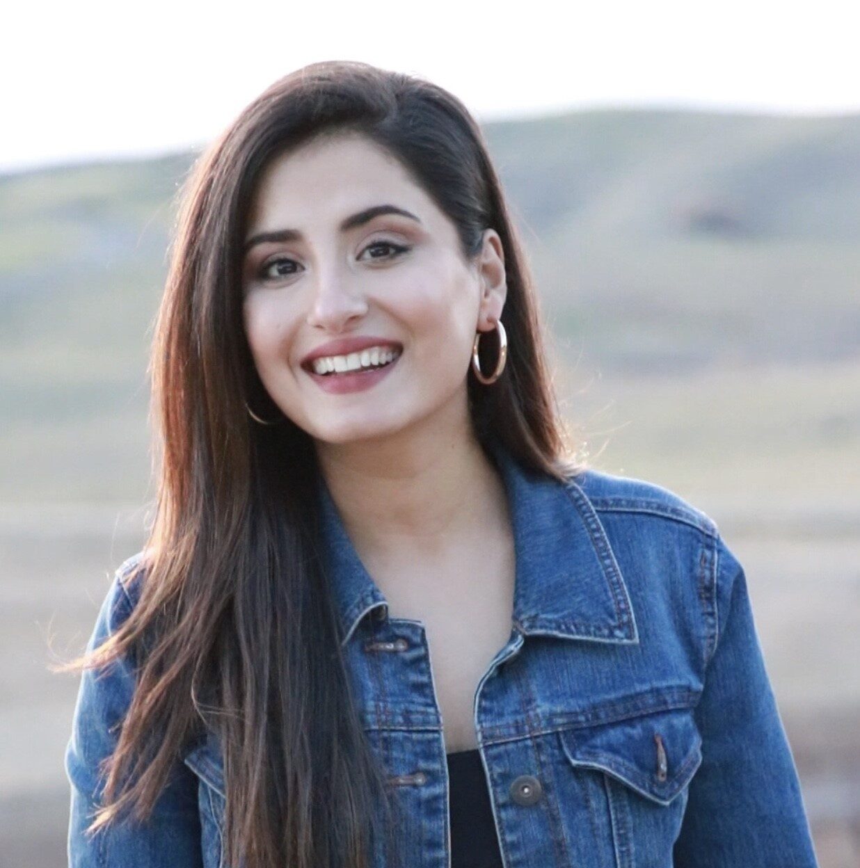 Sadaf wears a denim jacket and stands in front of a hilly background.