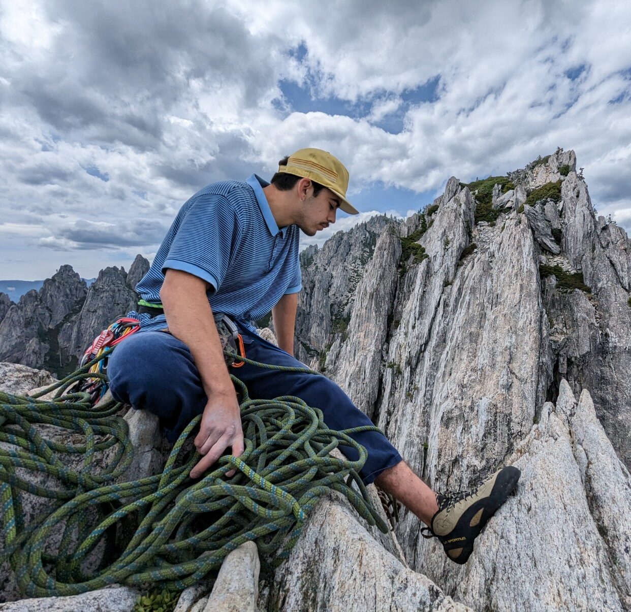 Photo of Jake atop a mountain of grey stone. He is wearing a blue shirt and tan hat. There is green rock climbing rope in the foreground.
