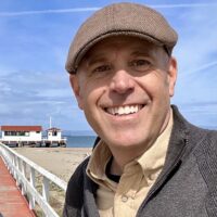 Brad wears a brown hat and grey jacket and stands in front of a pierhouse along the bay.