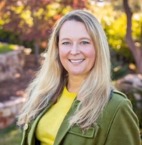 Monika Krach poses in front of fall foliage and wears a green jacket and yellow shirt.