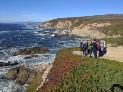 A group of Beach Watch volunteers stand on a rocky cliff overlooking the Pacific Ocean within Greater Farallones National Marine Sanctuary.